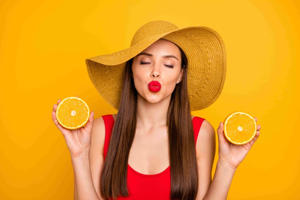 Attractive young woman looking at camera and covering eye with a fruit while standing against orange background - Beauty-lush-medspa