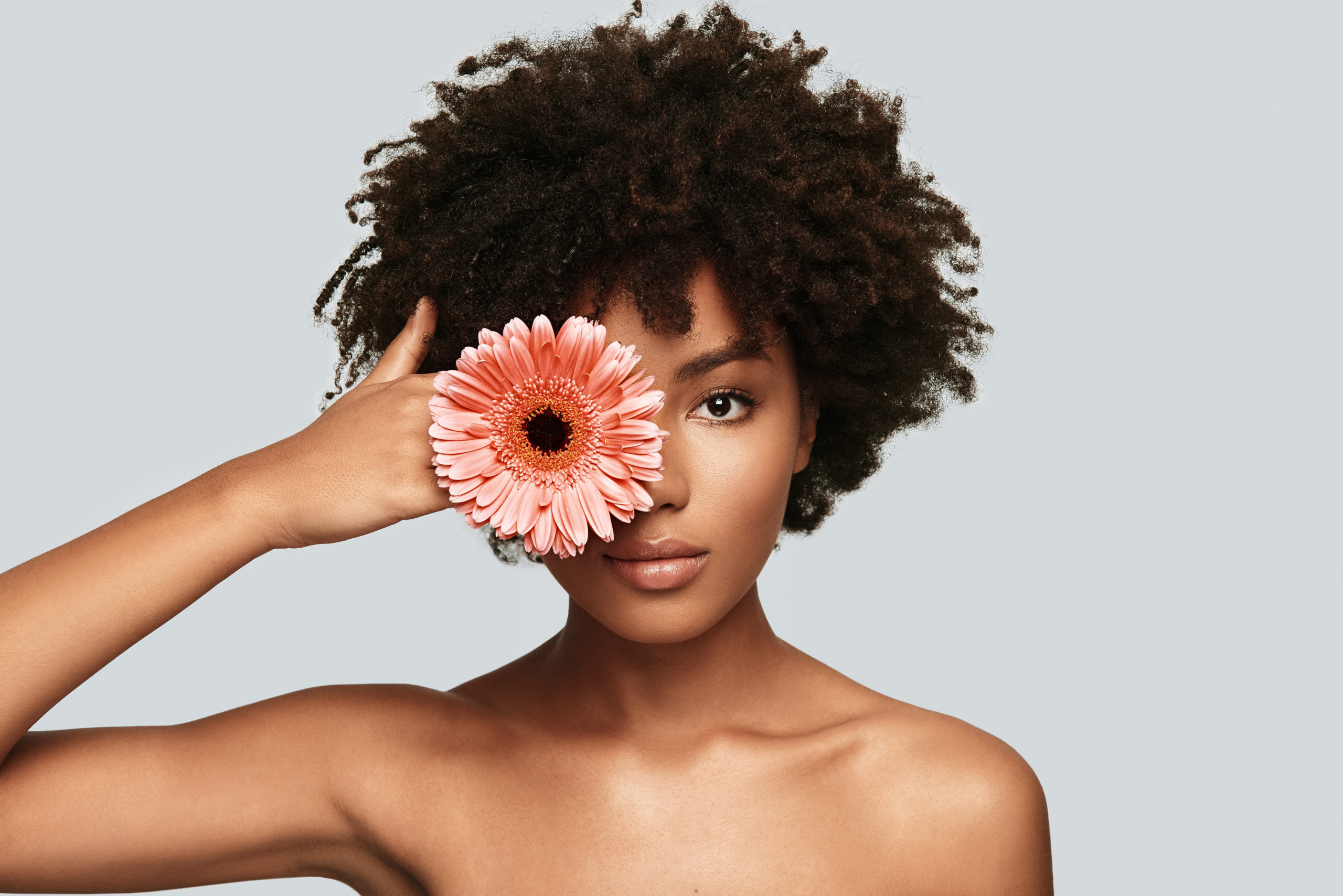 Beautiful as flower. Attractive young African woman looking at camera and covering eye with a flower while standing against grey background